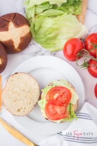 vertical photo of an open pretzel bun with lettuce and tomato fixings placed on the bottom half