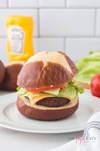 vertical photo of ninja foodi air fryer burger complete with burger patty, cheese, lettuce and tomato inside. On a white plate with fixings and mustard in the background