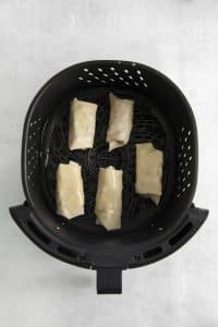 Top view photo of 5 rolled egg rolls, in the basket of the air fryer, ready to cook.