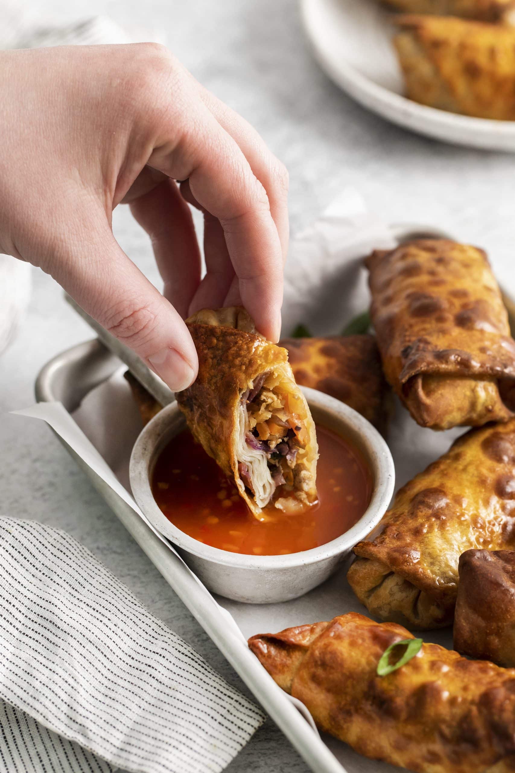 Photo of an Air Fryer Egg Roll, cut in half, and a hand dipping the egg roll in a dipping sauce in a small white bowl. 