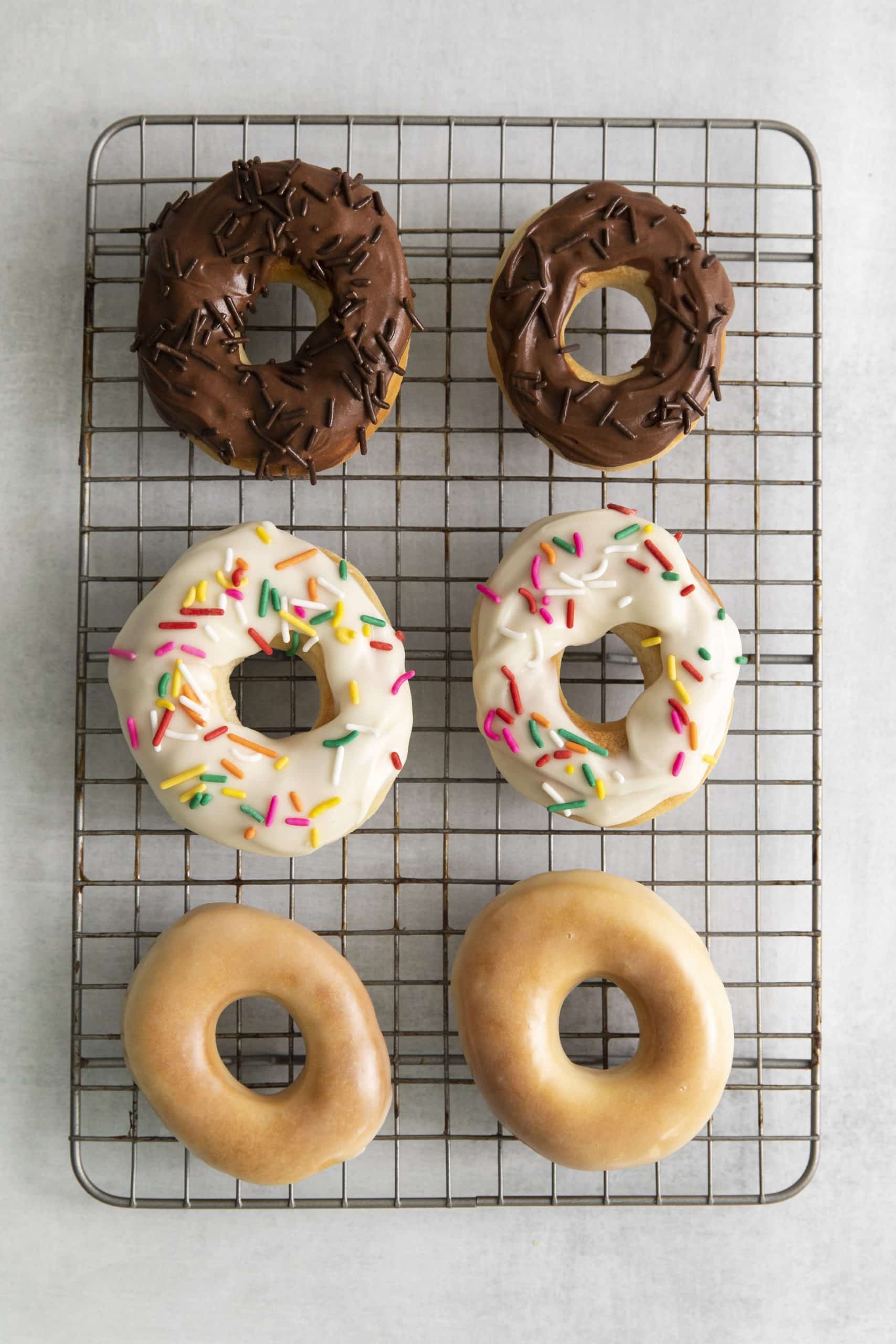 Top view photo of glazed and frosted Air Fryer Donuts on a cooling rack. Two are glazed, two have vanilla frosting with sprinkles, and two have chocolate frosting with chocolate sprinkles.