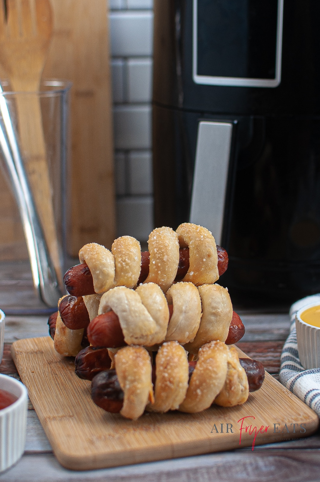 vertical photo of Air Fryer Pretzel Dogs stacked on a cutting board with an air fryer, utensils in a jar and dips surrounding them, all on a wooden surface