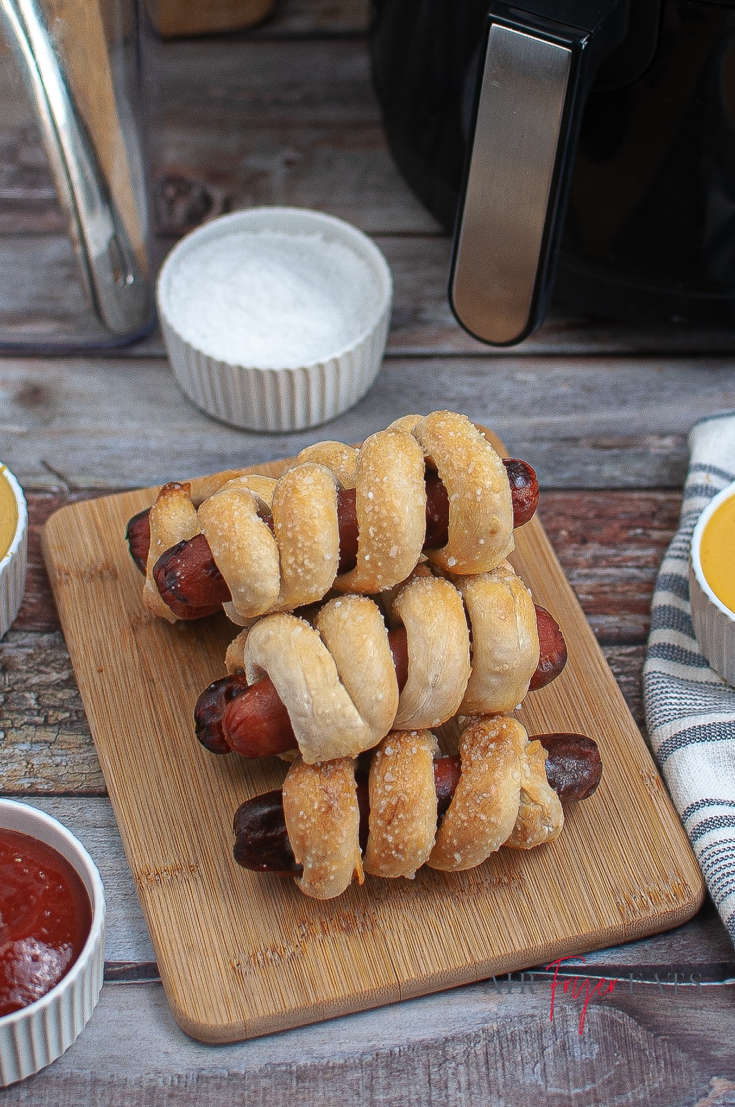 vertical photo of 5 air fryer pretzel dogs on a cutting board with an air fryer, a selection of dips and a ramekin with salt in the background. All on a wooden surface