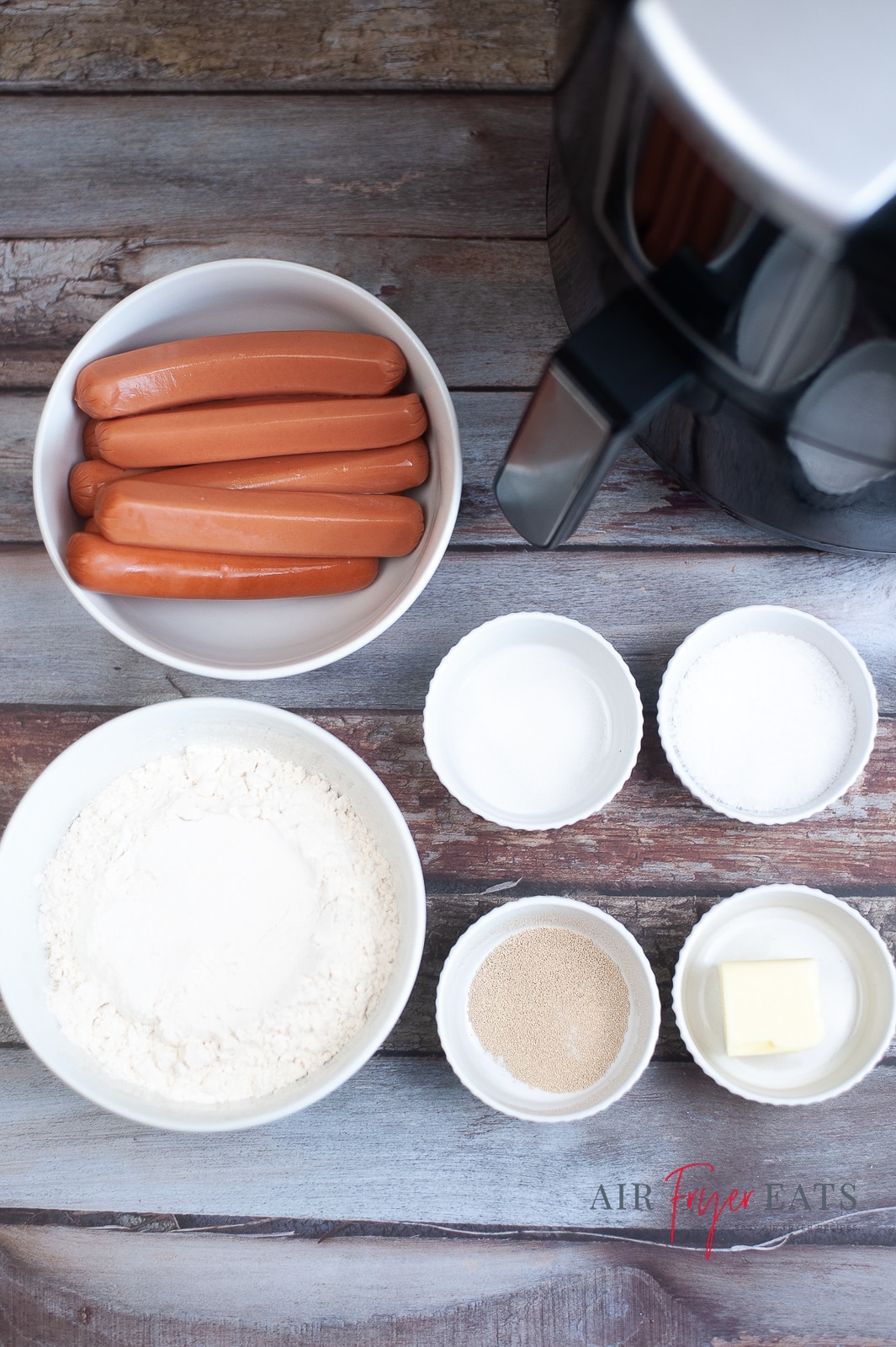vertical photo showing the ingredients for Air Fryer Pretzel Dogs: hotdogs, flour, butter, water, yeast, sea salt and sugar with an air fryer in the background, all on a wooden surface