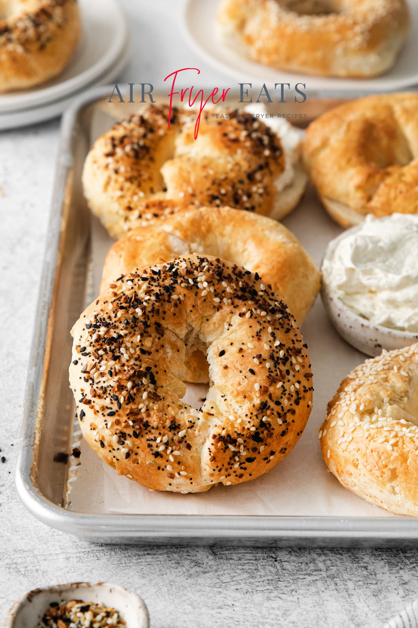 Photo of multiple air fryer bagels on a baking sheet with a small white bowl of cream cheese on the sheet.