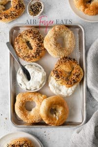 Top view photo of 5 air fryer bagels, on a baking sheet, next to a small bowl of cream cheese. One of the bagels is sliced open and has cream cheese spread on the inside.