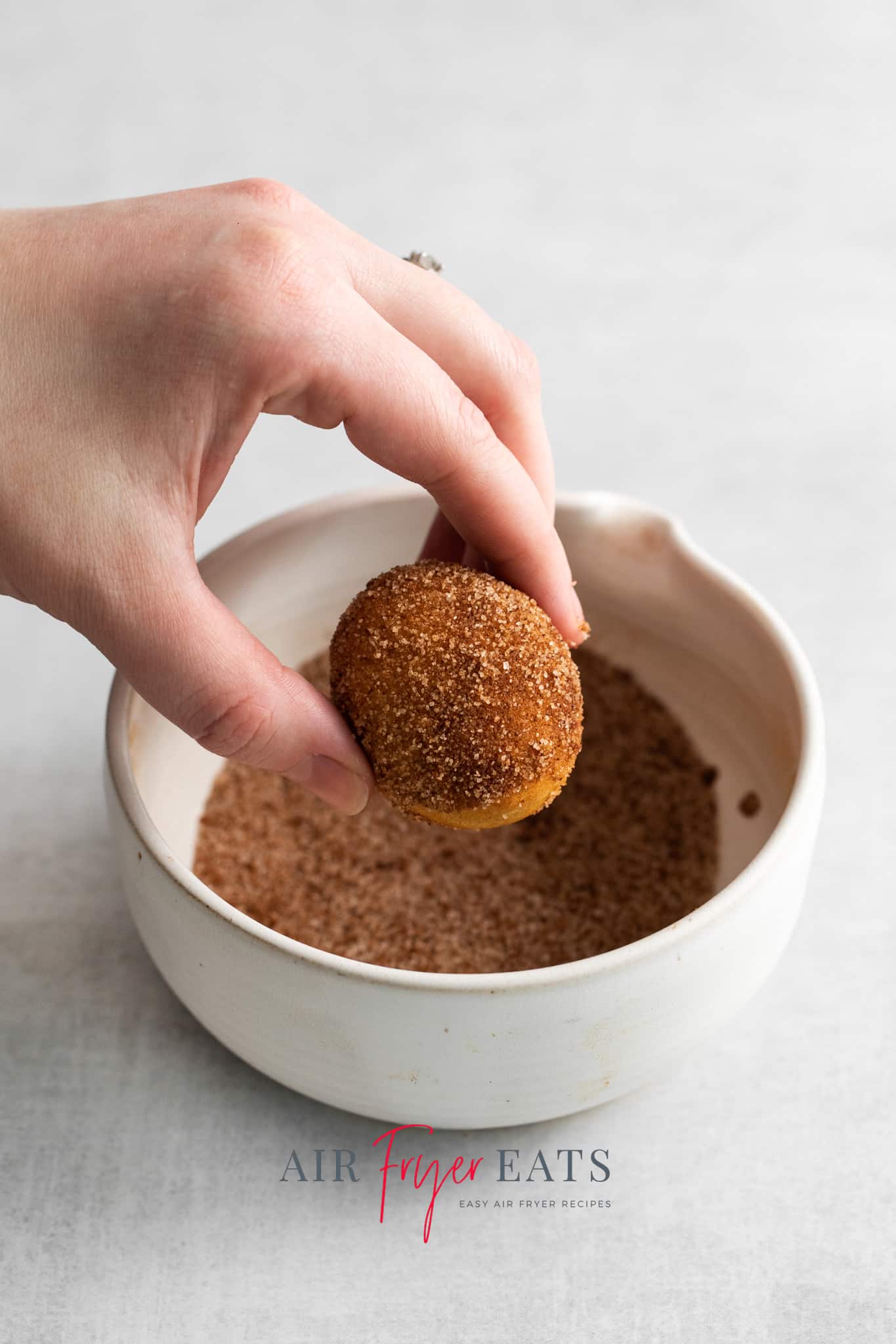 Photo of a hand dipping an Air Fryer Donut Hole into a small white bowl filled with a cinnamon-sugar topping. 