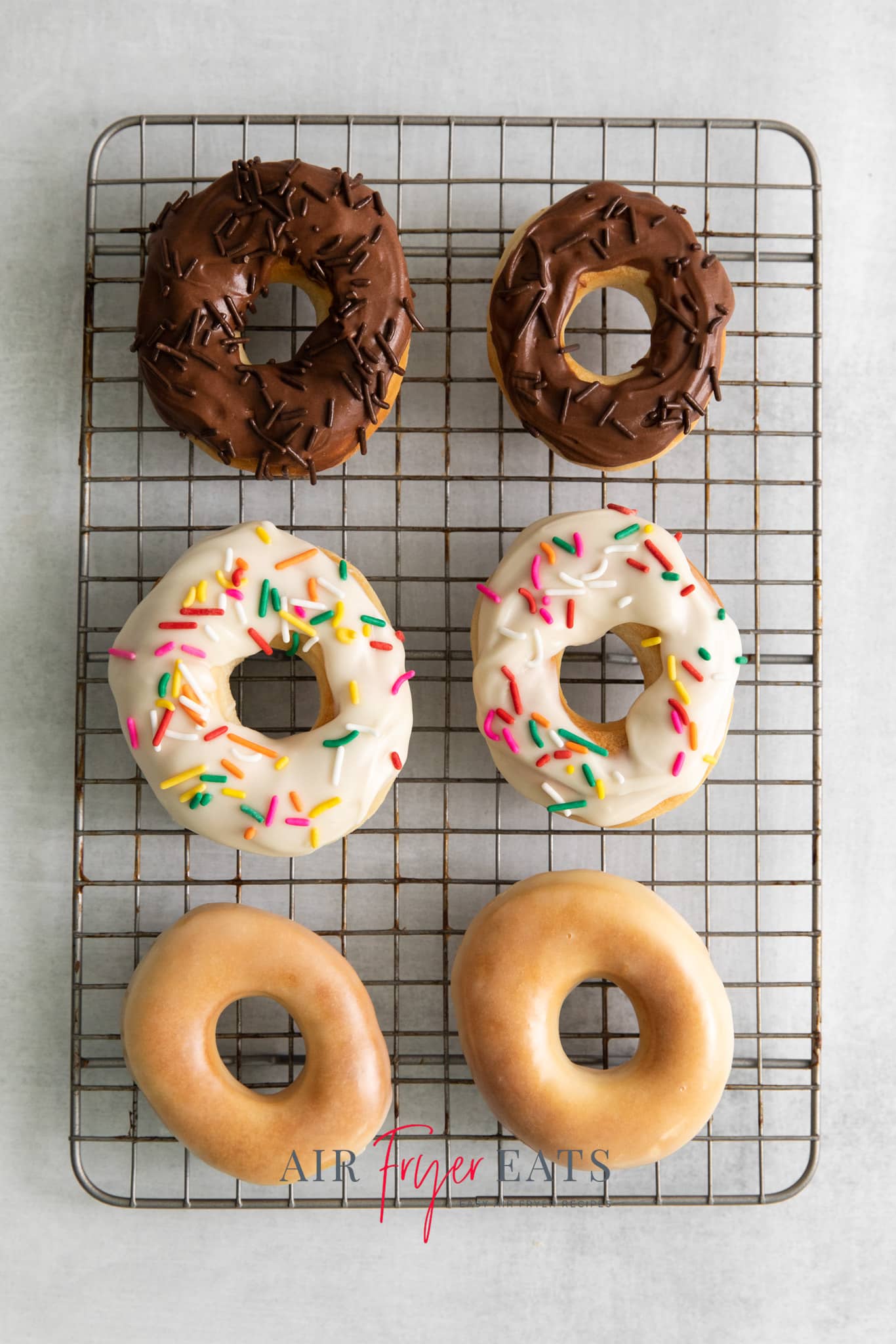 Top view photo of six air fryer doughnuts on a cooling rack. Two are glazed, two have vanilla frosting with colorful sprinkles, and two have chocolate frosting with chocolate sprinkles.