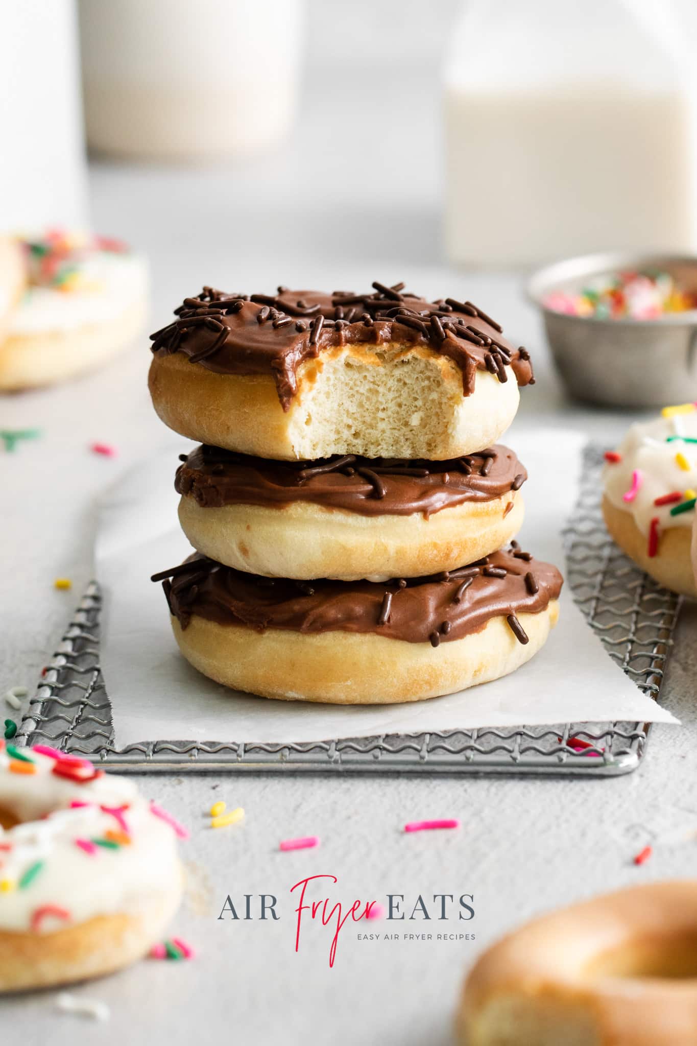 Photo of a stack of three air fryer doughnuts with chocolate frosting and chocolate sprinkles, on a cooling rack covered in parchment paper. 