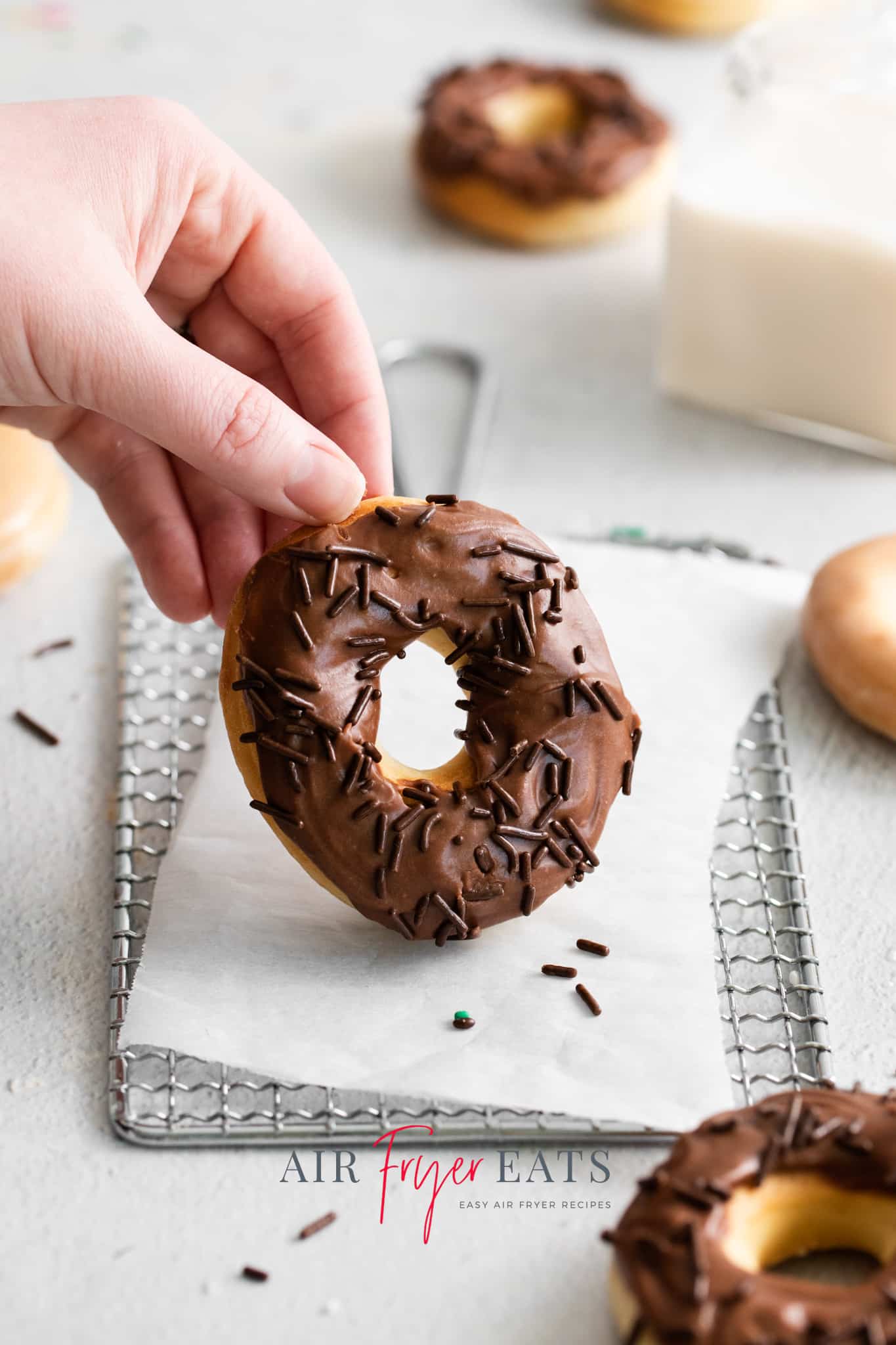Photo of a hand lifting an air fryer doughnut covered in chocolate frosting and chocolate sprinkles, on a parchment-lined cooling rack.