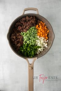 Top view photo of a frying pan with ground meat, carrots, onions, bell pepper, and peas, ready to cook together until soft.