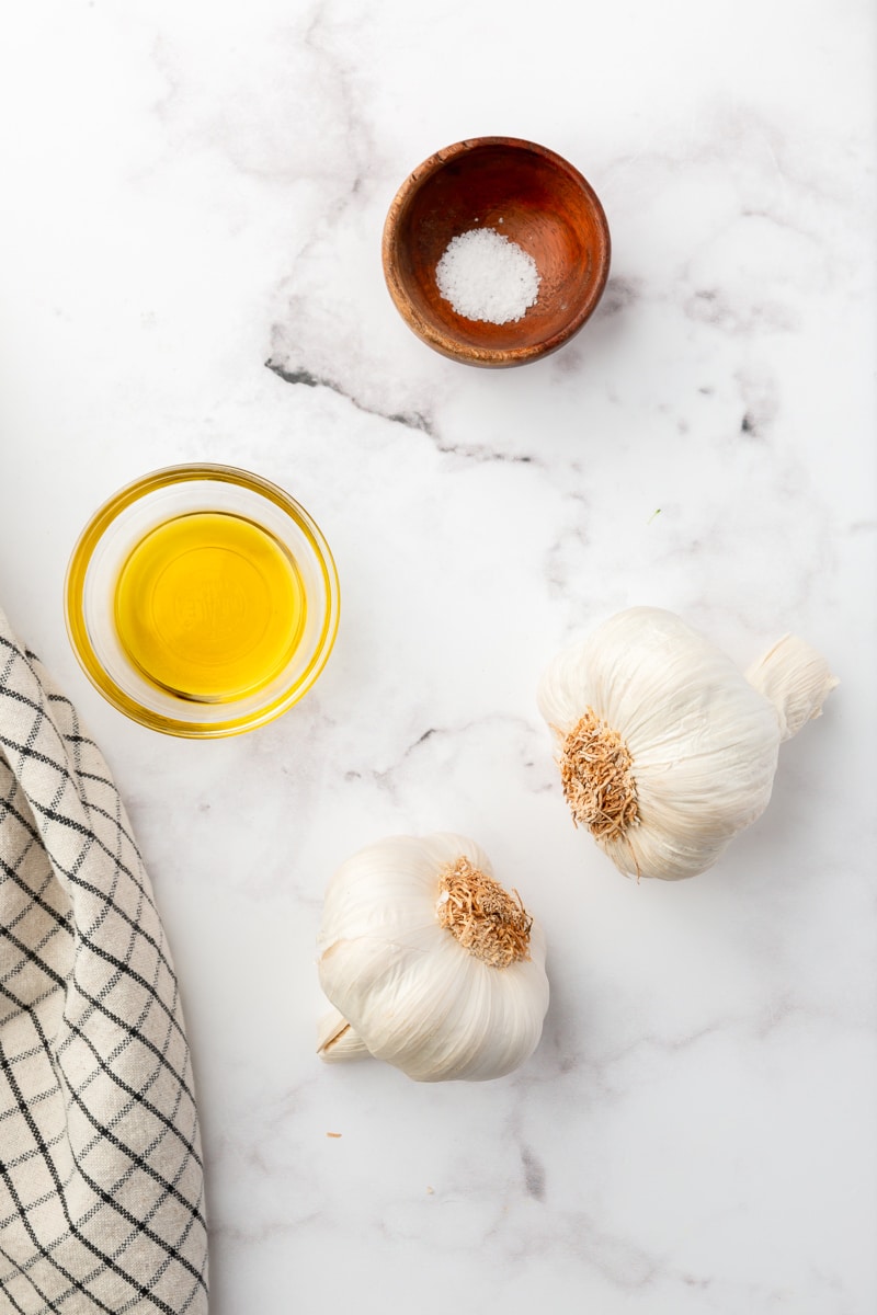 large garlic heads, olive oil, and kosher salt, on a marble countertop. 
