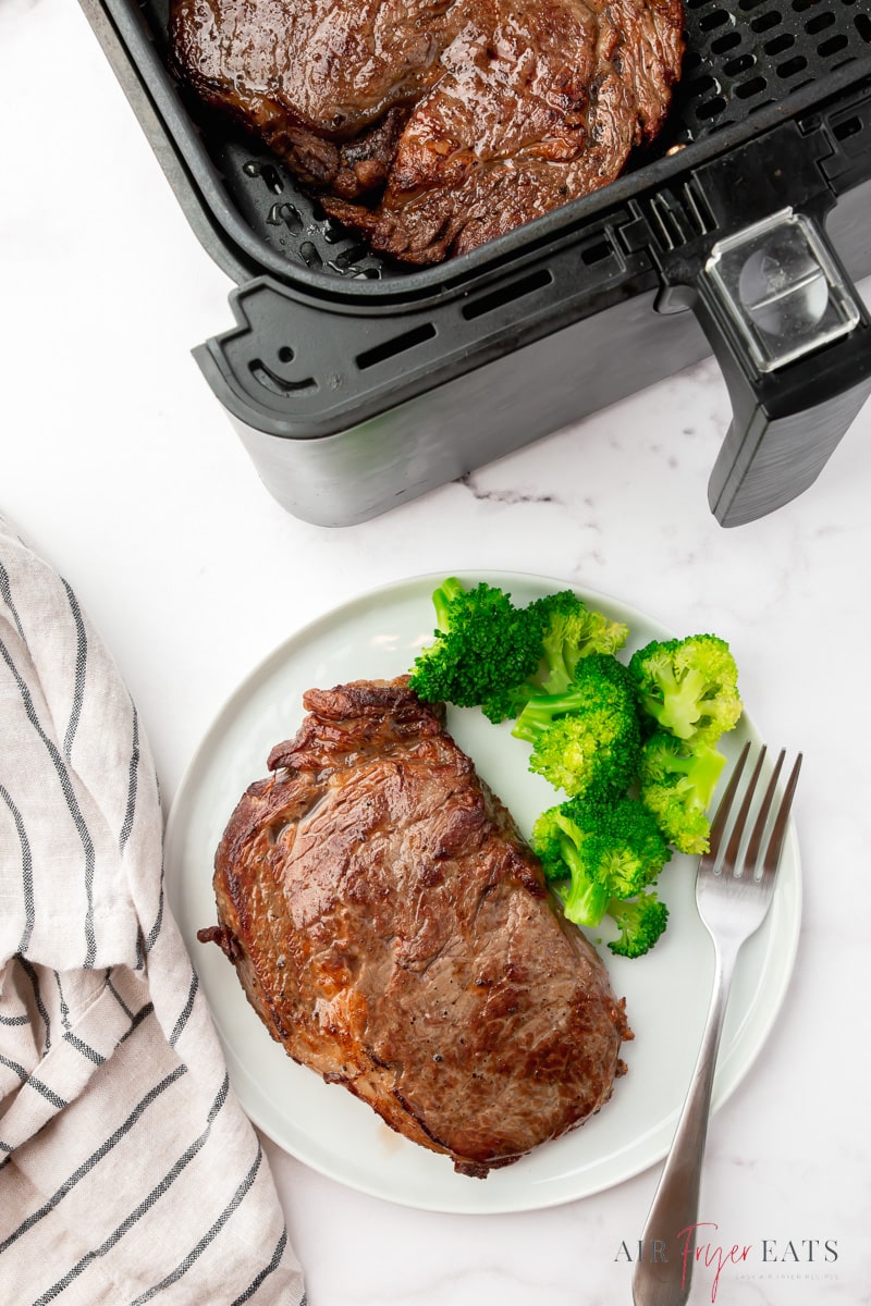 a plate of steak and broccoli, next to an air fryer basket with a steak in it.