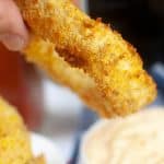 vertical photo of a close up of a air fryer cheesy onion ring held between thumb and forefinger with a plate of onion rings and a bowl of mayonnaise in the background