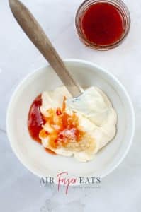 vertical photo showing mayonnaise, sweet chili sauce and minced garlic in a white bowl with a spatula. a bowl of sweet chili sauce is in the background. All on a white marble surface.