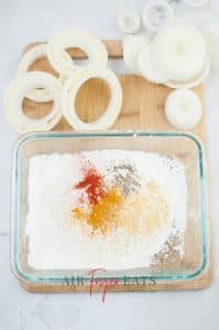 vertical photo showing cornstarch, flour and spices in a bowl, outside the bowl is cheese-stuffed onion rings all on a wooden board