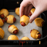 Photo of a hand holding an Air Fryer Pigs in a Blanket over the basket of the air fryer.