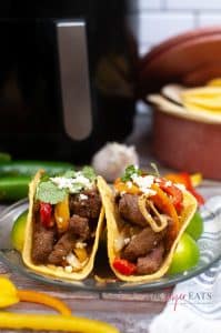 vertical photo of 2 fajita-filled tortillas on a glass plate with bell pepper strips in the foreground and bell peppers, white onion and tortillas out of focus in the background
