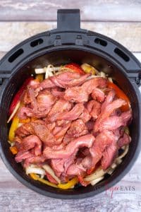 vertical photo of sliced and seasoned bell peppers, onion and beef strips in the air fryer basket
