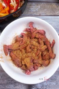 vertical photo of beef strips mixed with fajita seasoning in a bowl