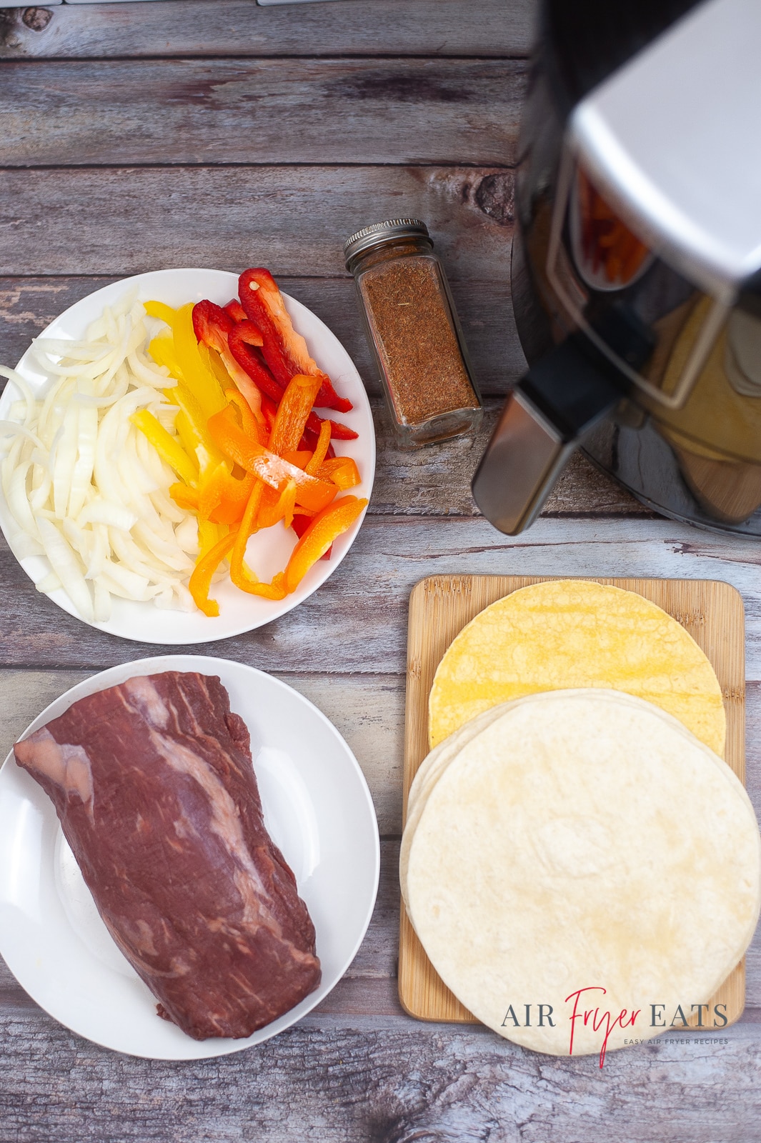 vertical photo of the ingredients for air fryer beef tenderloin fajitas: sliced bell peppers and white onion, beef tenderloin, fajita seasoning and tortillas. On a wooden surface with an air fryer in the top corner