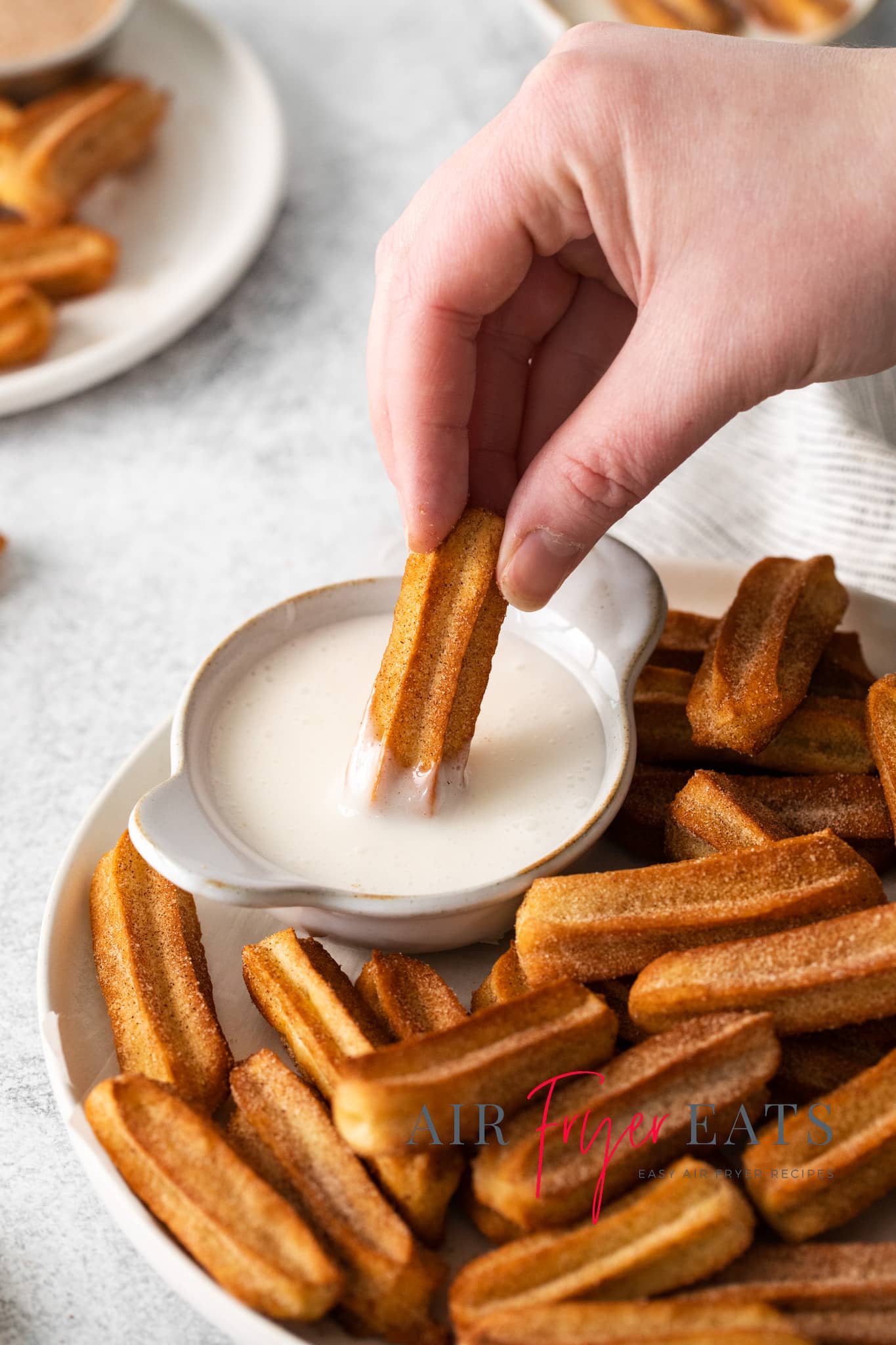 Photo of a hand dipping an Air Fryer Churro into a small bowl of powdered sugar glaze. 