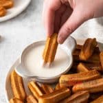Photo of a hand dipping an air fryer churro in a sweet dipping sauce. The sauce is in a small white bowl on a plate filled with air fryer churros.