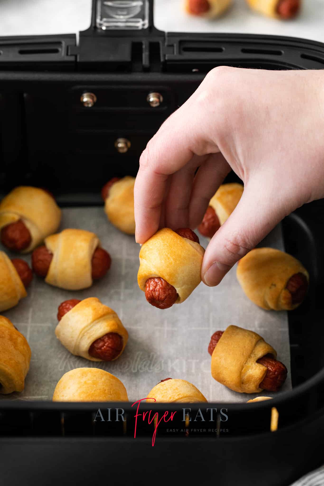 Photo of a hand holding an Air Fryer Pigs in a Blanket over the basket of the air fryer.