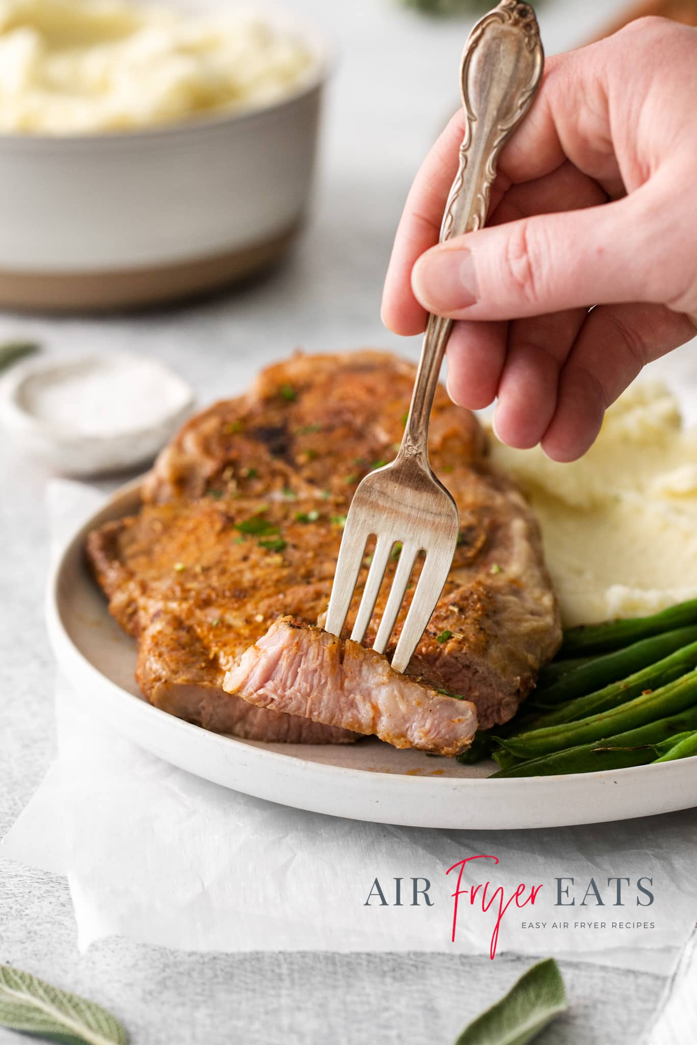Photo of a hand holding a fork with a bite of Air Fryer Pork Steak, ready to eat. It's on a plate with the rest of the pork steak, plus mashed potatoes and green beans. 
