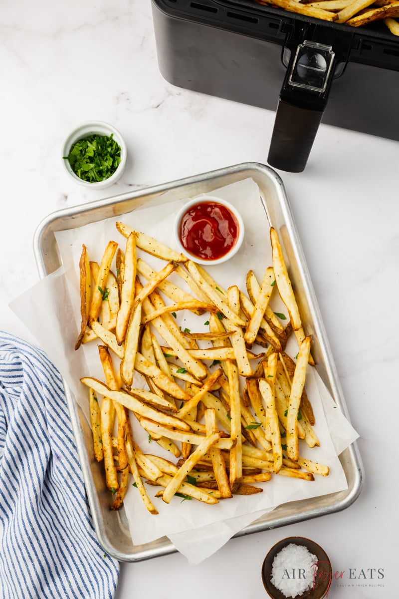 a small sheet pan lined with parchment paper, holding freshly cooked french fries and a small bowl of ketchup. An air fryer basket is next to the tray, as well as a small dish of chopped parsley. 