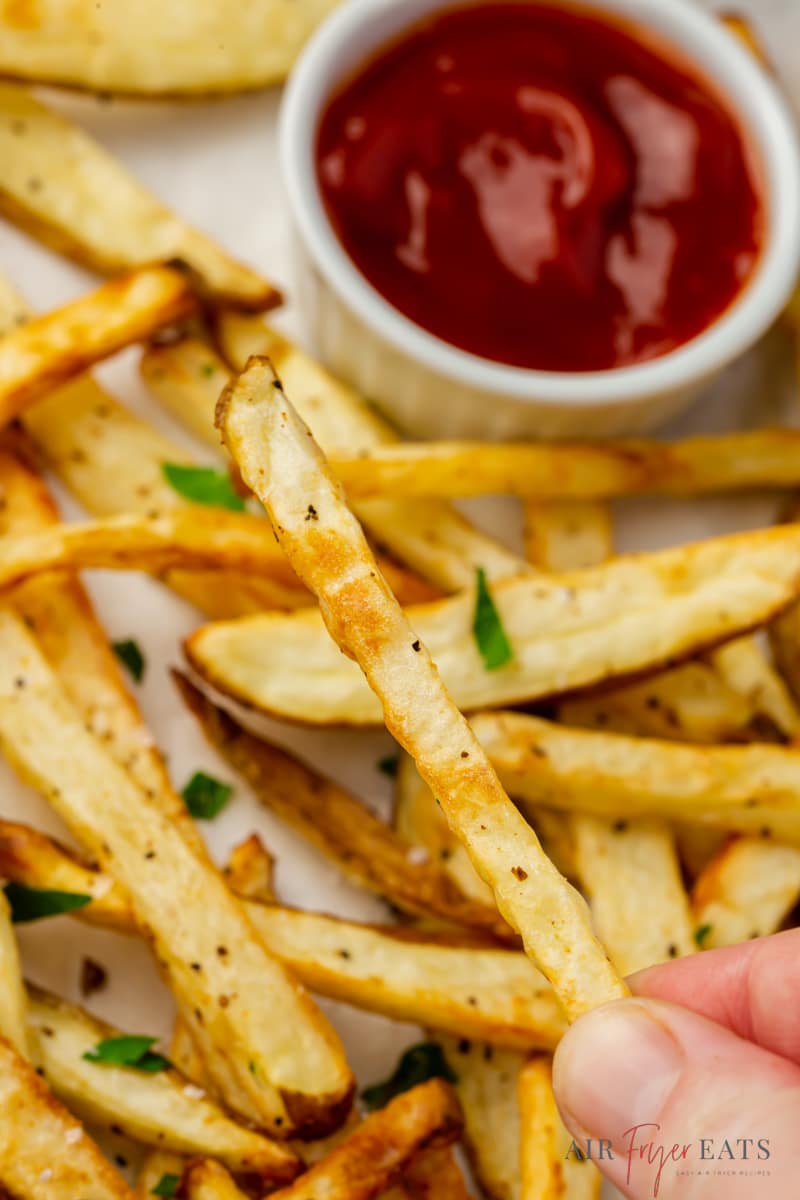 A hand is holding a crispy air fryer french fry over a plate of fries with a side of ketchup.