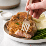 Photo of a hand holding a fork with a bite of Air Fryer Pork Steak, ready to eat. It's on a plate with the rest of the pork steak, plus mashed potatoes and green beans.