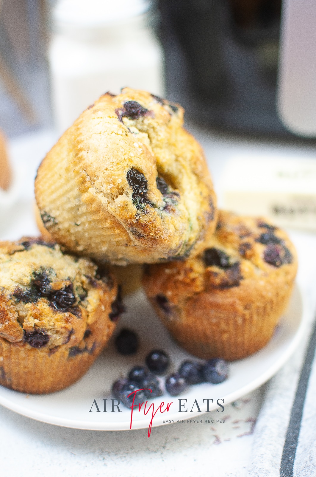 vertical photo showing a plate with four air fryer blueberry muffins and a few loose blueberries. On a white surface