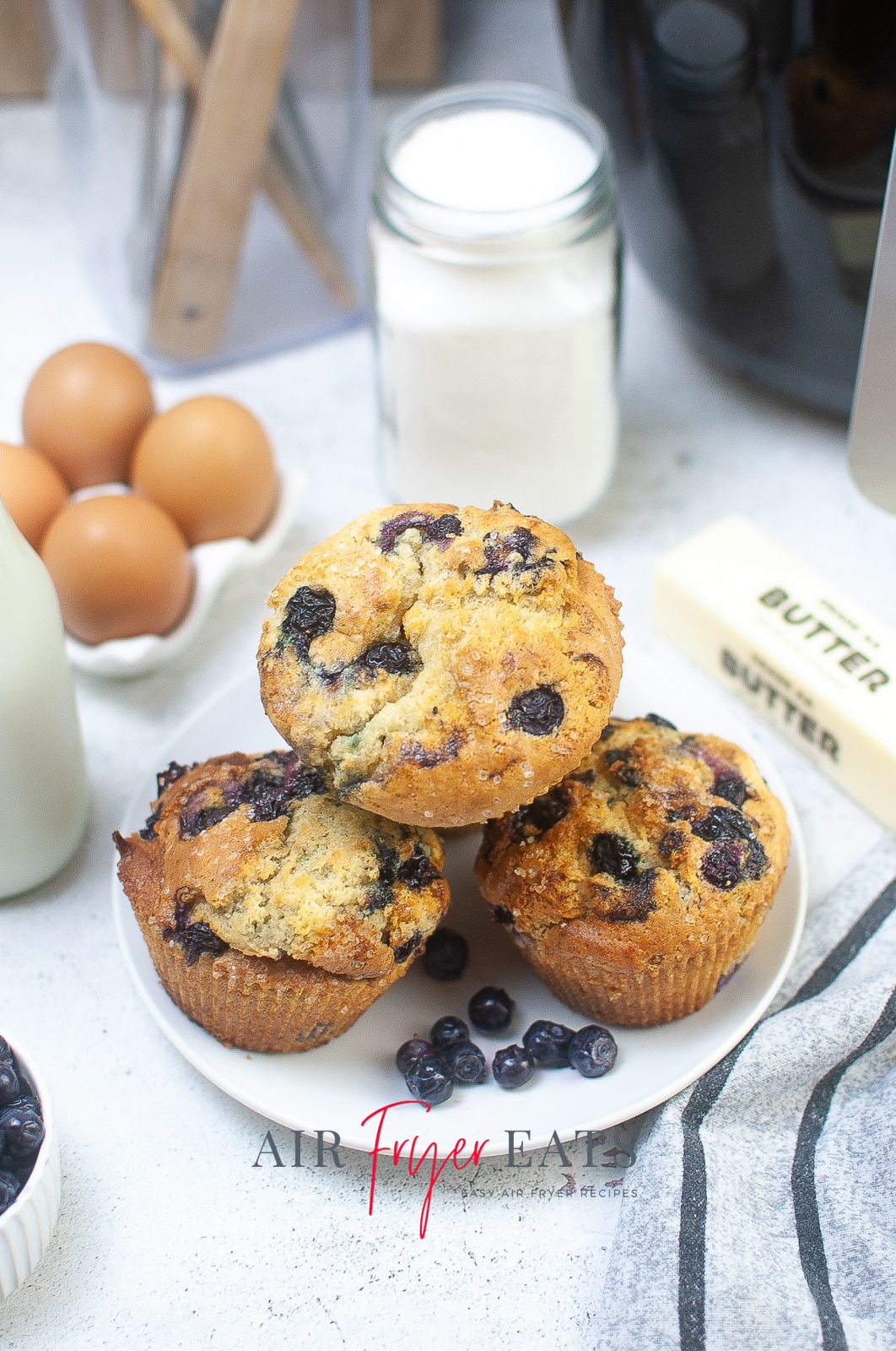 vertical photo showing a white plate with four air fryer blueberry muffins, with sugar, butter, eggs, milk and blueberries around the plate. All on a white surface