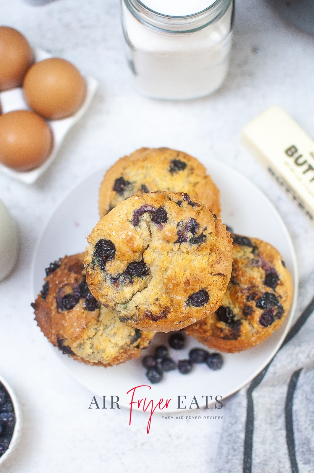 Vertical photo of an overhead shot of a plate with 4 blueberry muffins and a few loose blueberries. Eggs, sugar, butter and blueberries are around the plate, all on a white surface