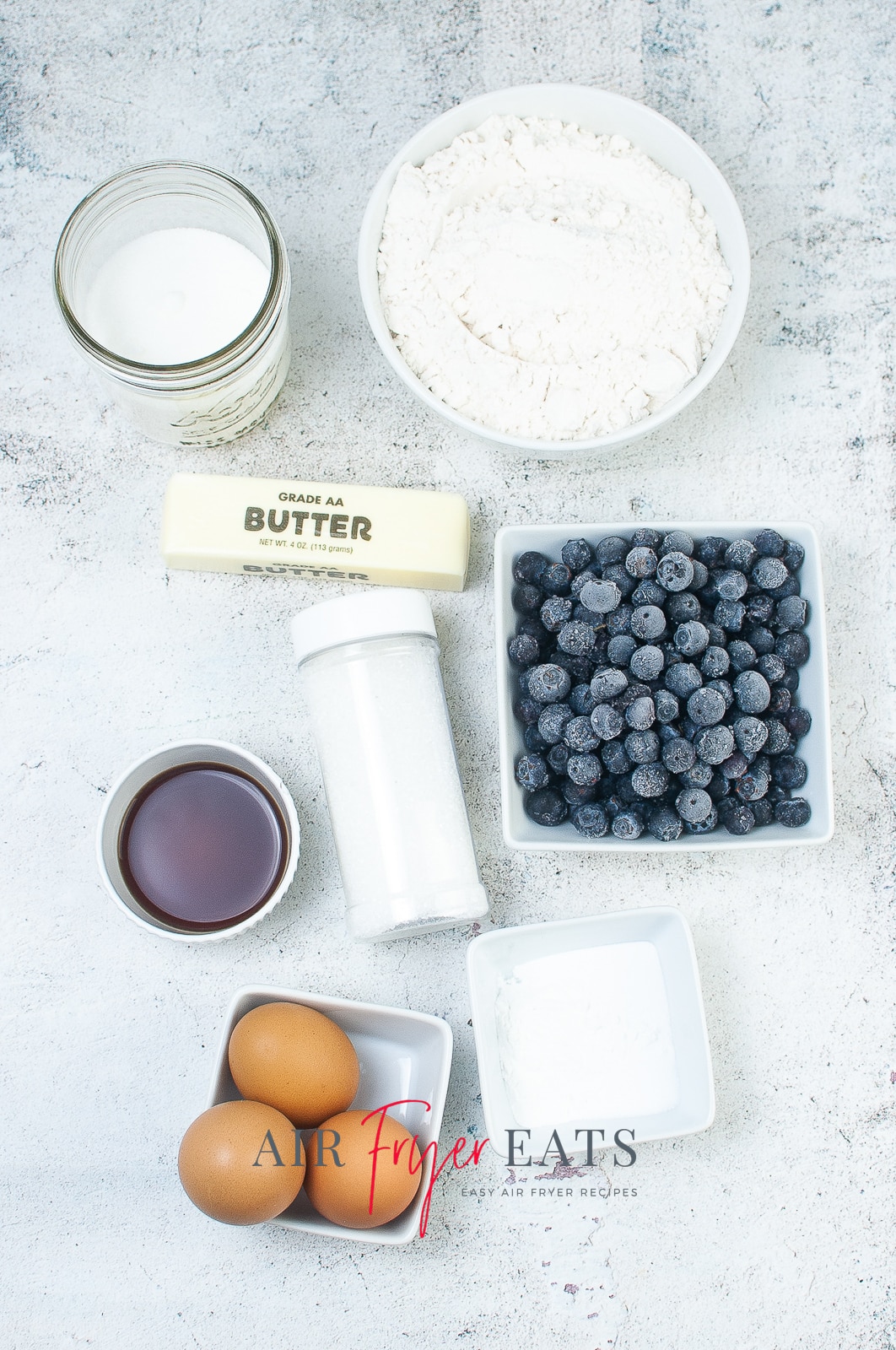 vertical photo showing ingredients for air fryer blueberry muffins: flour, baking powder and baking soda, salt, butter, sugar, eggs, milk, vanilla, blueberries