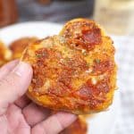 vertical photo showing a held heart shaped pepperoni pizza in the foreground with a plate of pizzas in the background
