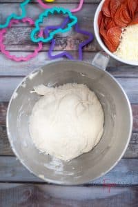 Vertical photo showing kneaded dough in a bowl with cookie cutters and pepperonis and cheese in the background, all on a wooden surface