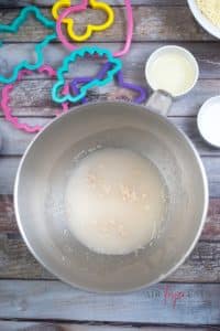vertical photo showing a bowl with water and yeast with cookie cutters in the background, all on a wooden surface