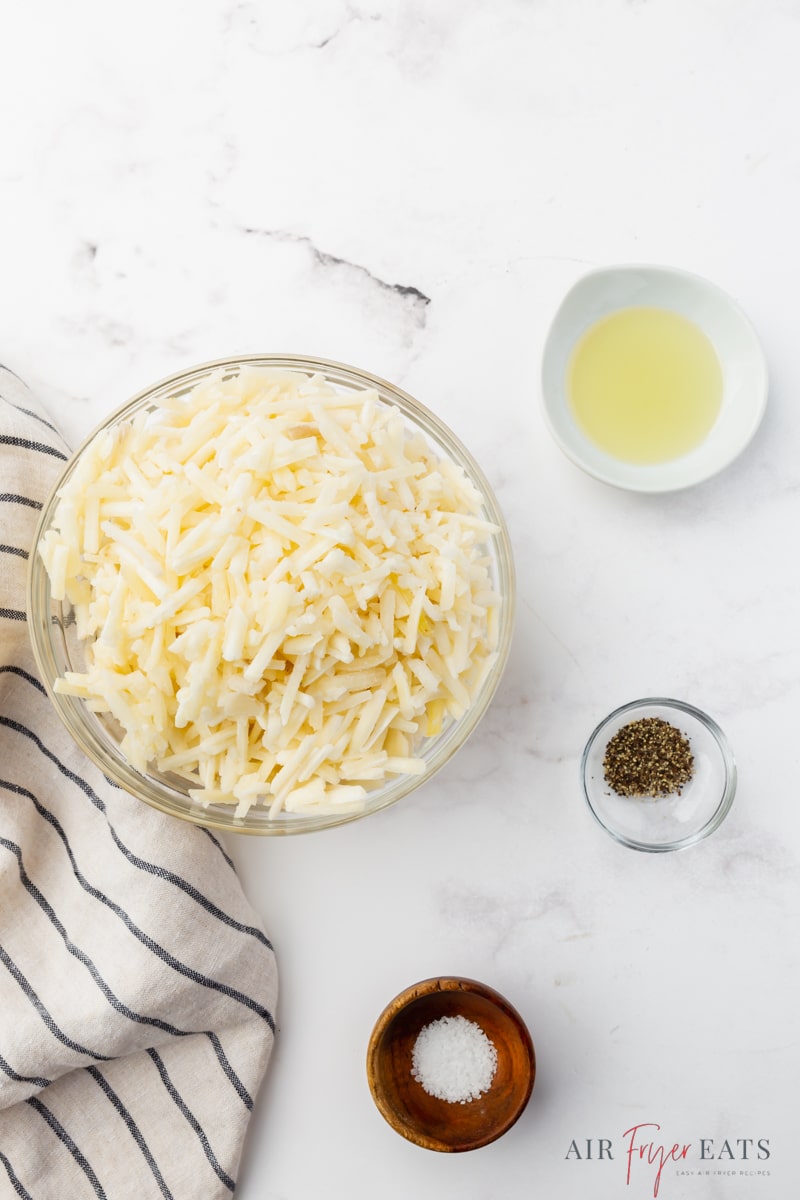 a bowl of frozen shredded potatoes next to small bowls of oil, salt, and pepper. 