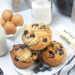 square photo showing four air fryer blueberry muffins on a white plate with some loose blueberries on the plate. Other ingredients are behind the plate