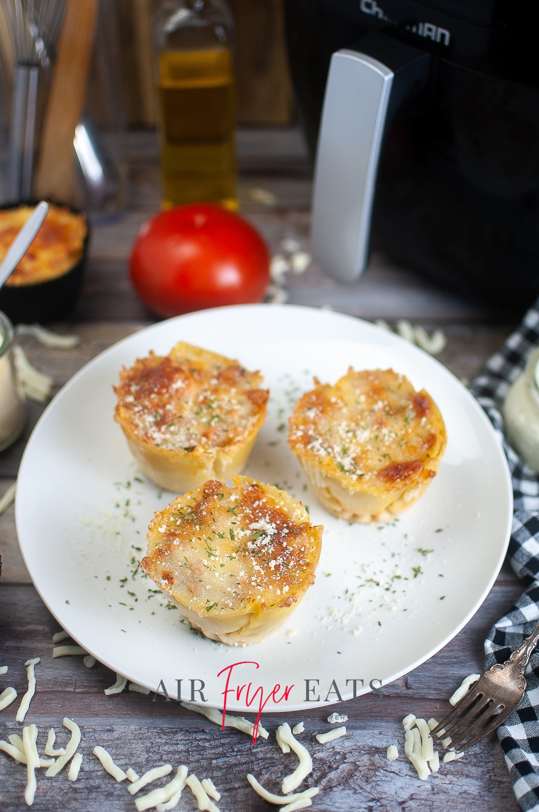 vertical photo showing 3 air fryer lasagna cups on a white plate with an air fryer and other ingredients in the background. All on a wooden surface