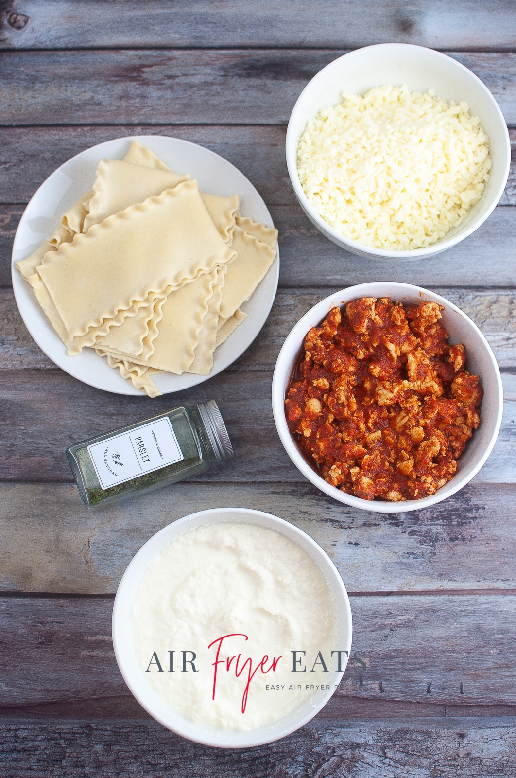 vertical photo showing the ingredients for air fryer lasagna cups: lasagna, meat sauce, ricotta cheese, mozzarella, parsley. All on a wooden surface