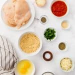 The ingredients needed for making air fryer chicken parmesan, including chicken cutlets, breadcrumbs, eggs, and cheese, measured into bowls and arranged on a marble countertop.