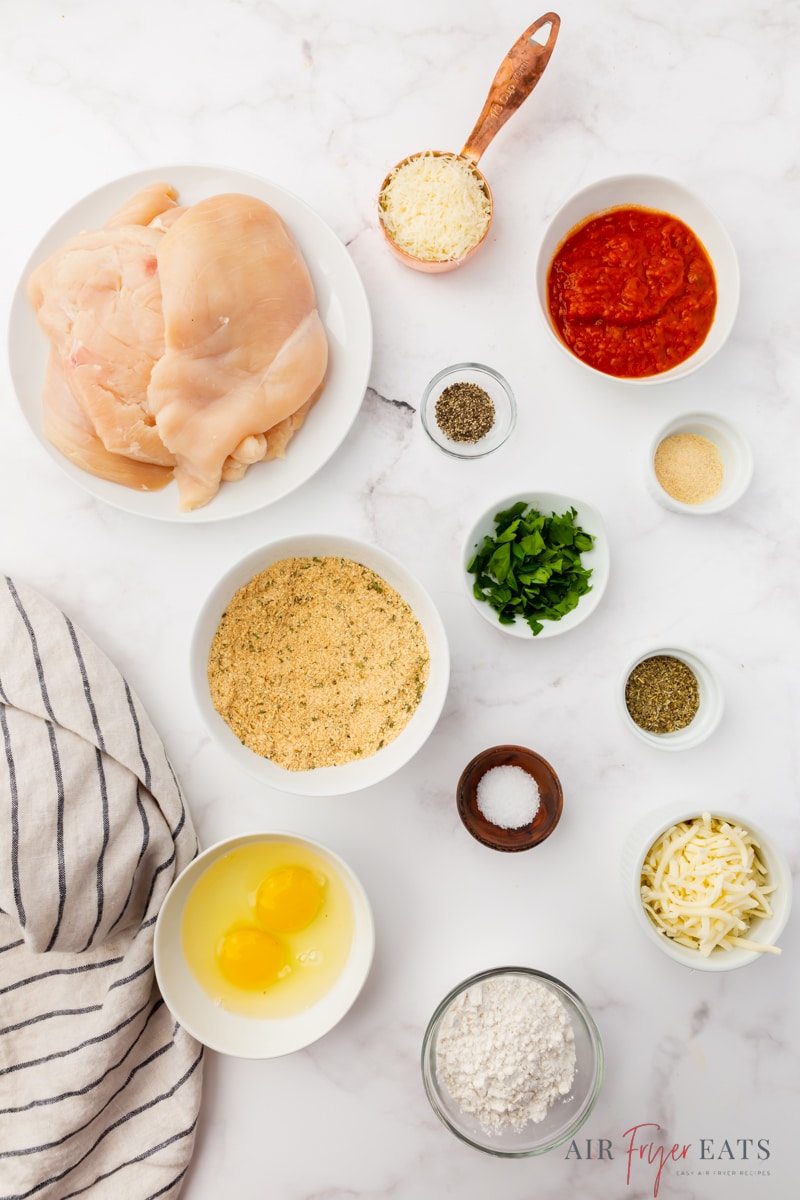 The ingredients needed for making air fryer chicken parmesan, including chicken cutlets, breadcrumbs, eggs, and cheese, measured into bowls and arranged on a marble countertop.