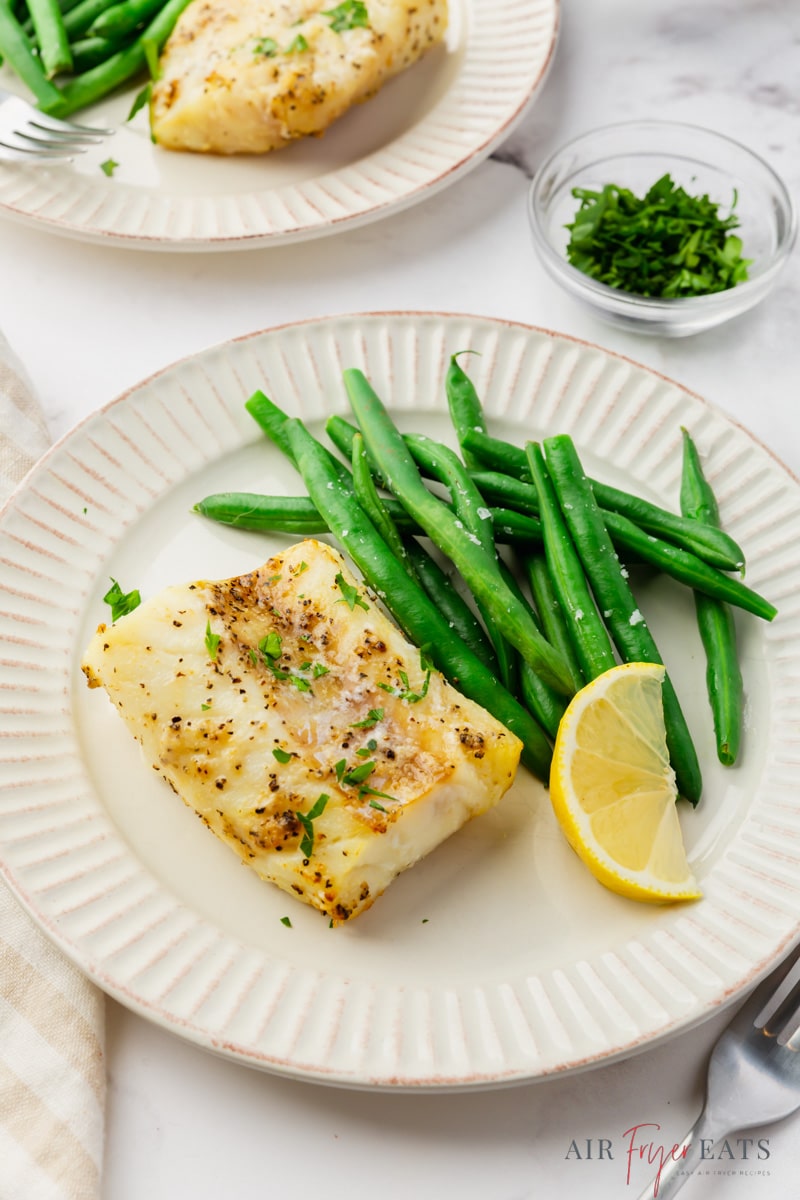 a round dinner plate with a serving of air fryer cod, green beans, and a lemon wedge. 