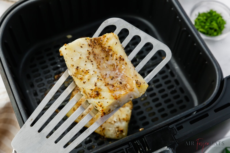A fish spatula holding up a cod fillet over an air fryer basket. 