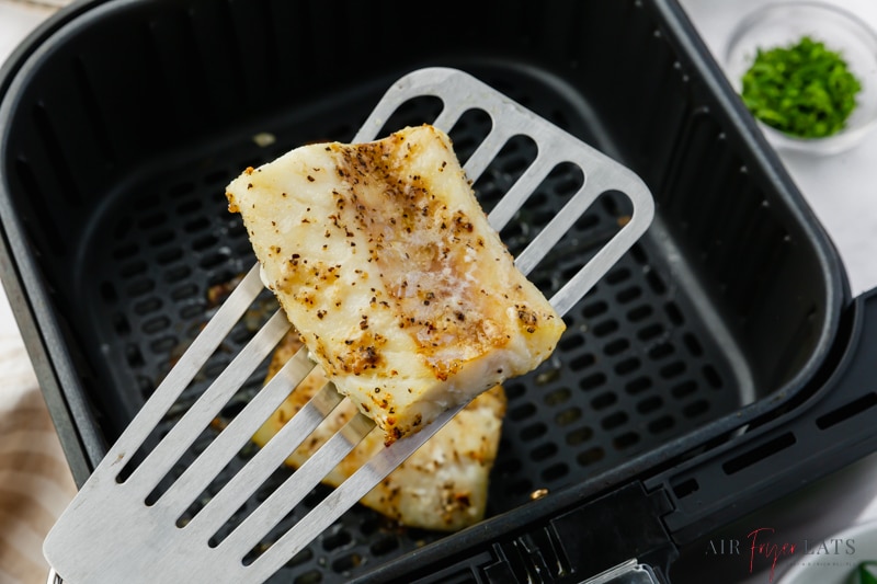 A fish spatula holding up a cod fillet over an air fryer basket.