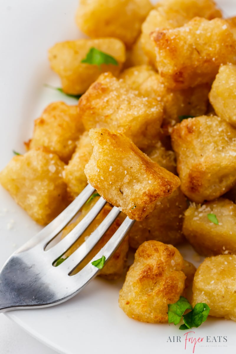 Photo of a fork with a golden brown air fried cauliflower gnocchi at the end of the fork, ready to enjoy. There is a plate of gnocchi in the background. 