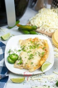 vertical photo showing a square white plate with air fryer cheese enchiladas, a lime wedge, a spoonful of sour cream and a couple of slices of green chiles. Lime wedges, shredded cheese on a board, tortillas, an air fryer and green chiles surround the plate